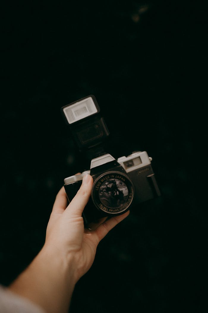 A vintage film camera with flash is held in a hand against a dark background, showcasing classic photography.